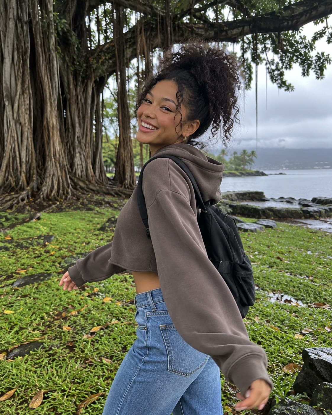 A smiling young woman with curly hair in a high puff, a cropped brown hoodie, jeans and a backpack twirls under giant banyan trees by Hilo Bay on an overcast day: Rockstarr Media model casting in Hilo, Hawaiʻi Island