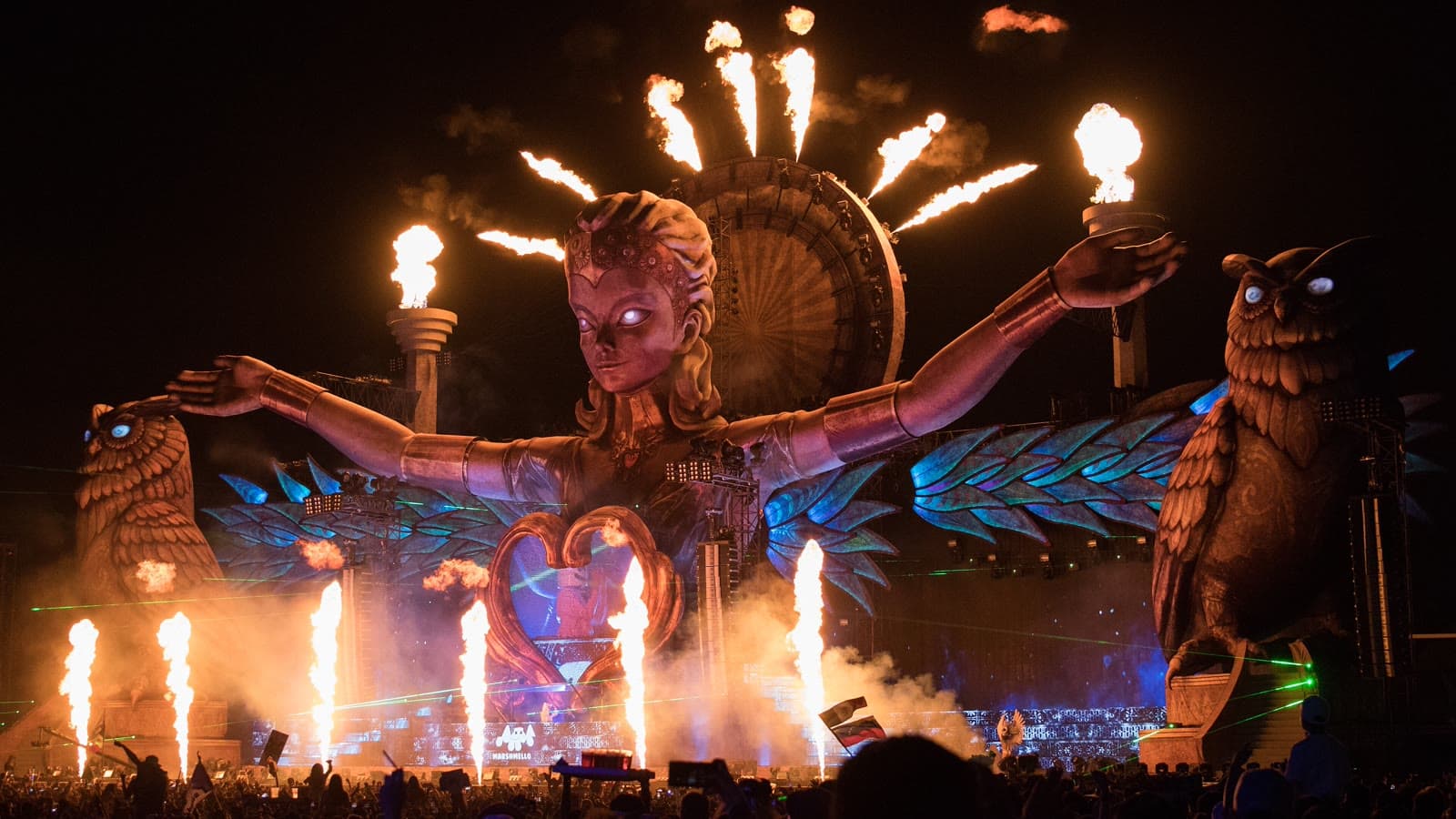 Flames shoot up around a giant goddess figure and owl sculptures on an EDC mainstage at night as a huge crowd watches, the festival behind Hotel EDC Orlando 2026